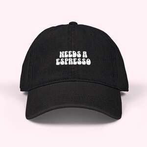 May include: Black baseball cap with the white text "NEEDS A ESPRESSO" embroidered on the front. The cap has a curved brim and an adjustable strap at the back. The hat is made of a soft, durable fabric.