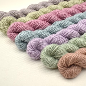 May include: A collection of colorful yarn skeins in various pastel shades, including lavender, pink, and green. The yarn is arranged in diagonal rows, showcasing the different colors and textures. The skeins are tightly wound, suggesting a high-quality fiber for crafting.