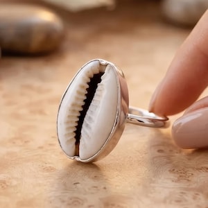 Può includere: Un anello in argento con una conchiglia di ciprea bianca incastonata in una montatura in metallo liscio. La conchiglia presenta una distinta apertura centrale con bordi seghettati e il gambo dell'anello è in metallo color argento lucido.