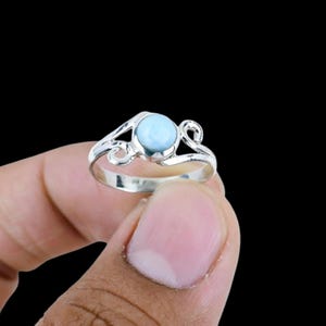 May include: A silver ring featuring a round, light blue cabochon stone set in the center. The band of the ring is crafted from polished silver and displays decorative, swirling metalwork patterns that extend from the central stone setting.