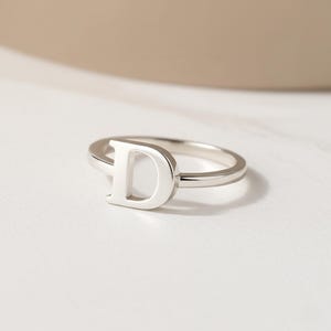 Könnte beinhalten: Ein polierter Ring in Silberoptik mit einem markanten Großbuchstaben D als Herzstück. Die Ringschiene ist glatt und kreisförmig und weist eine saubere, metallische Oberfläche auf, die das Licht reflektiert. Der Buchstabe D ist in den Ring integriert.