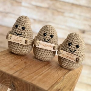 May include: Three crocheted potato-shaped figures with smiling faces and small wooden signs. The signs read "You are loved", "You got this!", and "You are Spudtacular!". The figures are brown and sit on a wooden surface.