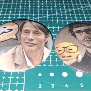 Puede incluir: Dos botones redondos con imágenes de hombres y una moneda. Un botón presenta a un hombre con un bocadillo, el otro a un hombre con un emoji con gafas. La moneda es dorada y plateada.