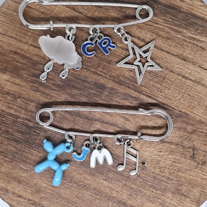 May include: Two silver safety pins with decorative charms. One pin has a cloud, blue letters "CR", and a star charm. The other pin has a blue balloon animal, the letter "J", a "W", and a musical note charm. The pins are on a wooden surface.