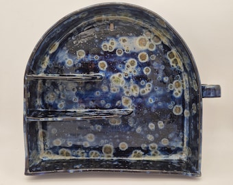 Può includere: Un portasapone in ceramica fatto a mano con una glassa blu scuro e un motivo maculato. Il piatto ha una base rettangolare con due ripiani e una parte superiore curva, e un piccolo manico laterale. Il piatto è largo circa 25 cm.
