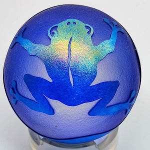 May include: A round, blue glass paperweight with a frog design. The frog is in a lighter blue and iridescent colours, with its arms and legs outstretched. The paperweight is on a clear stand.