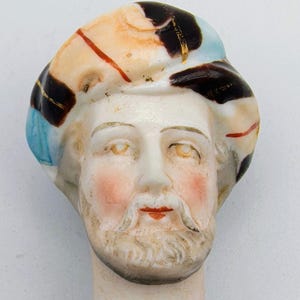 May include: A vintage porcelain doll head with a painted face, beard, and hair. The doll head has a light skin tone with rosy cheeks and red lips. The hair is a mix of colors, including brown, orange, and blue. The doll head is mounted on a white stem.