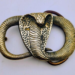 May include: A metallic gold-toned belt buckle featuring a raised, detailed cobra snake design. The snake is coiled into an oval shape with its hood flared and head centred. The surface displays a textured scale pattern throughout the body.