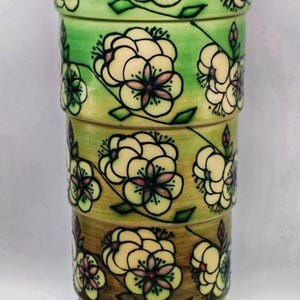 Puede incluir: Un jarrón de cerámica apilado con un diseño floral. El jarrón está compuesto por varias secciones cilíndricas, cada una adornada con flores color crema con centros rosas y hojas verdes sobre un degradado de tonos verdes y marrones.