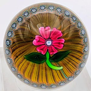 May include: A spherical glass paperweight featuring a central pink flower with a white centre containing the letter P, a green stem and leaves, set against a brown and yellow radial background, surrounded by a border of small blue and white floral canes.