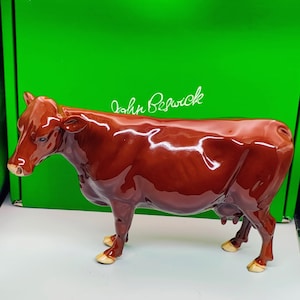 May include: A glossy, brown ceramic cow figurine. The cow has a detailed design with a shiny finish. The figurine is standing on a white surface, with a green background behind it. The text "John Beswick" is visible on the green background.