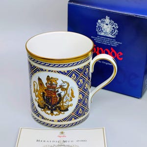 May include: A Spode Heraldic mug with a gold rim and handle, displaying a royal crest and blue and gold geometric patterns. The mug is presented with a blue box and a certificate. The mug is a commemorative item.
