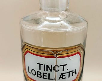 May include: A clear glass apothecary bottle with a stopper. The bottle has a rectangular label with the text "Tinct. Lobel. Æth." in black lettering, framed by gold and red lines. The bottle is empty.