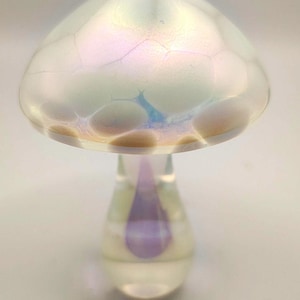 Pode incluir: Um cogumelo decorativo de vidro com um chapéu iridescente e um caule transparente. O chapéu tem um padrão rachado com tons de azul e dourado. O caule apresenta um detalhe roxo em forma de gota. Um item de decoração único.