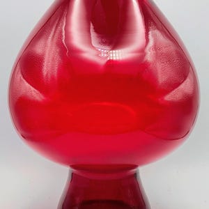 Könnte beinhalten: Eine leuchtend rote Glasvase mit bauchigem Körper und ausgestelltem Fuß. Die Vase hat eine glatte, glänzende Oberfläche und eine breite Öffnung oben. Die Gesamtform erinnert an einen Tropfen. Die Vase ist ein dekorativer Artikel.