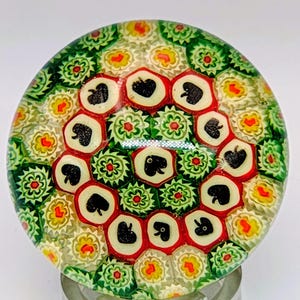 Puede incluir: Un pisapapeles redondo de cristal con un diseño floral colorido. El pisapapeles presenta un anillo central de diseños negros en forma de corazón rodeados de patrones florales rojos y verdes. Los bordes exteriores están llenos de patrones florales amarillos, verdes y blancos.