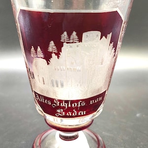 May include: A clear glass goblet with a red base and rim, featuring a detailed white etched design of a castle and trees. The text "Altes Schloss von Baden" is displayed in red lettering.