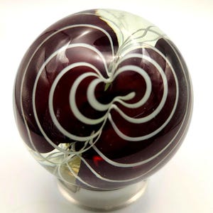 Può includere: Una sfera decorativa in vetro di colore bordeaux intenso, con linee bianche concentriche e vorticose. La sfera poggia su una base color argento, creando un contrasto sorprendente. Il design ricorda un occhio stilizzato o un motivo complesso.