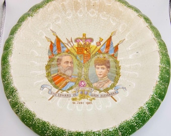 Op de afbeelding: Een decoratieve keramische plaat met een groene geschulpte rand. Het midden toont portretten van koning Edward VII en koningin Alexandra, met vlaggen en een kroon. De tekst luidt "Coronation 26 June 1902."