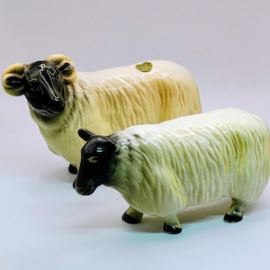 Puede incluir: Dos figuras de ovejas de cerámica. Las ovejas tienen caras y patas negras, con lana de color crema. Una oveja es más grande y está de pie, mientras que la otra es más pequeña y está en posición de pastoreo. Una pegatina dorada está en la oveja más grande.