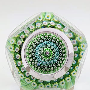 Könnte beinhalten: Ein Briefbeschwerer aus klarem Glas mit einem komplexen Millefiori-Design aus konzentrischen Kreisen mit grünen, weißen und blauen blumenartigen Mustern. Die Mitte zeigt einen kleinen violetten Punkt, umgeben von sternförmigen Glasstäben.