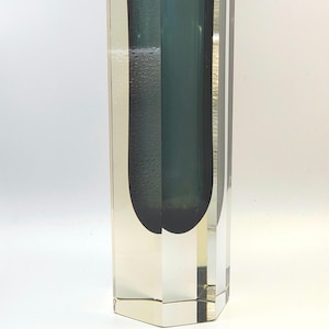 Puede incluir: Un jarrón alto de vidrio transparente con forma rectangular y un interior verde oscuro. El jarrón tiene una superficie texturizada en un lado y un acabado liso y pulido en el otro. La base es un diseño multifacético.