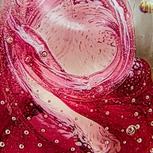 Può includere: Primo piano di un fermacarte in vetro con un design astratto a spirale. Il fermacarte presenta sfumature di rosso e rosa, con un vortice centrale e piccole bolle d'aria. Il vetro ha una finitura lucida.