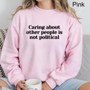 May include: A light pink sweatshirt with the text "Caring about other people is not political" in black. The sweatshirt is a solid color and has a crew neck.