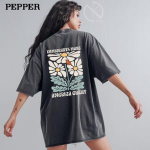 May include: Dark gray t-shirt with a daisy graphic and the text "IMMIGRANTS MAKE AMERICA GREAT". The graphic features white daisies with yellow centers and a butterfly. The shirt has short sleeves and a relaxed fit.