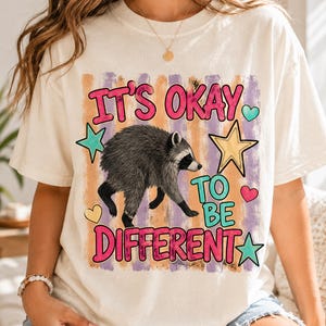 Pode incluir: Uma t-shirt de cor creme com um gráfico de um guaxinim ao centro. O texto IT'S OKAY TO BE DIFFERENT está escrito em rosa e turquesa. O design inclui riscas verticais cor de laranja e roxas, estrelas amarelas e turquesas e pequenos corações.