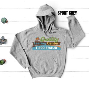 May include: Sport grey hoodie with a kangaroo pocket and drawstring hood. The front features the text "Quality Learing Center" with a graphic and the phone number "1-800-FRAUD". The hoodie is made of a soft, comfortable material.