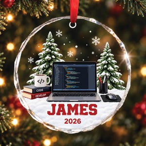May include: A circular glass ornament featuring a laptop displaying code, a mug with a code symbol, books labeled CODE and DEVELOP, and a mouse. The name JAMES and the year 2026 are printed in red text. The scene includes snowy pine trees.