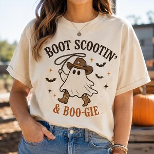 Może przedstawiać: Kremowy t-shirt z krótkim rękawem z grafiką ducha w kowbojskim kapeluszu i butach, trzymającego lasso. Napis "BOOT SCOOTIN' & BOO-GIE" wykonany jest brązowymi literami i otoczony małymi czarnymi nietoperzami oraz pomarańczowymi gwiazdkami.
