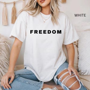 May include: White t-shirt with the word "FREEDOM" printed in black block letters. The word "WHITE" is printed in the upper right corner. The shirt has short sleeves and a crew neck.