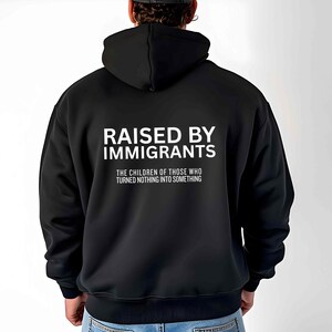 May include: Black hooded sweatshirt with the text "RAISED BY IMMIGRANTS" in white block letters. Below the text is the phrase "THE CHILDREN OF THOSE WHO TURNED NOTHING INTO SOMETHING". The sweatshirt is made of a soft material.
