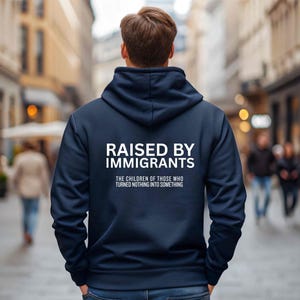 May include: Navy blue hoodie with the text "RAISED BY IMMIGRANTS" in white block letters. Below the text is the phrase "THE CHILDREN OF THOSE WHO TURNED NOTHING INTO SOMETHING". The person is standing in a city street.