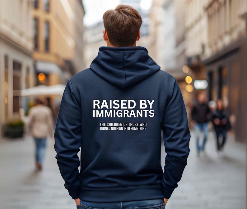 May include: Navy blue hoodie with the text "RAISED BY IMMIGRANTS" in white block letters. Below the text is the phrase "THE CHILDREN OF THOSE WHO TURNED NOTHING INTO SOMETHING". The person is standing in a city street.