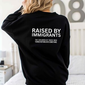 May include: Black sweatshirt with the text "RAISED BY IMMIGRANTS" in white block letters. Below the text is the phrase "THE CHILDREN OF THOSE WHO TURNED NOTHING INTO SOMETHING" in white. The sweatshirt is a long-sleeved crew neck.