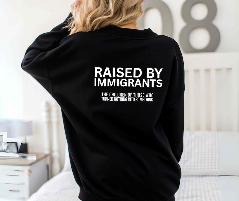 May include: Black sweatshirt with the text "RAISED BY IMMIGRANTS" in white block letters. Below the text is the phrase "THE CHILDREN OF THOSE WHO TURNED NOTHING INTO SOMETHING" in white. The sweatshirt is a long-sleeved crew neck.