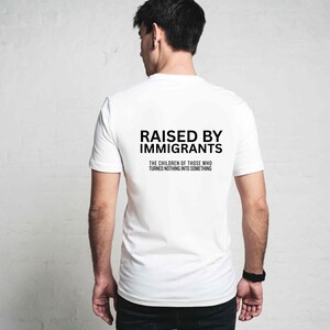 May include: White t-shirt with the text "RAISED BY IMMIGRANTS" in black, along with the phrase "THE CHILDREN OF THOSE WHO TURNED NOTHING INTO SOMETHING." The shirt is short-sleeved and appears to be made of cotton.