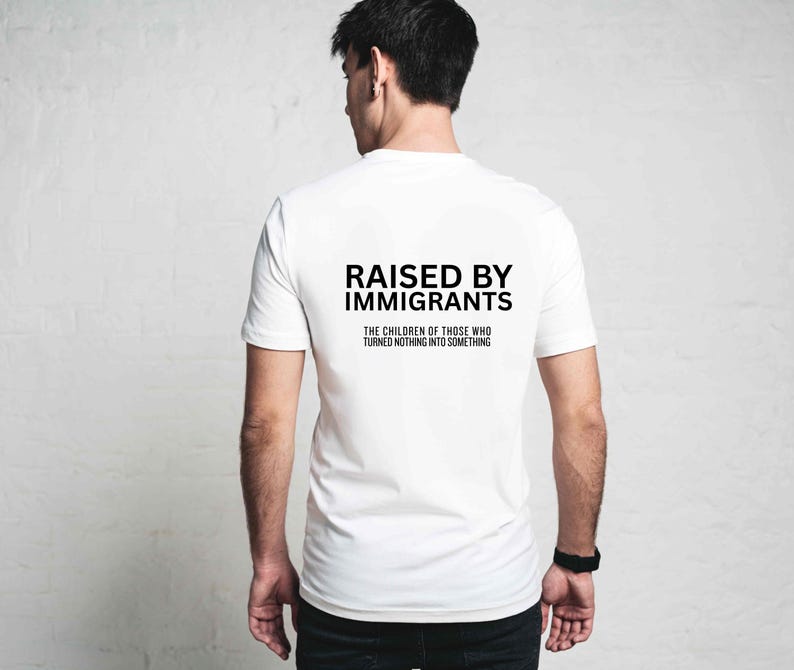 May include: White t-shirt with the text "RAISED BY IMMIGRANTS" in black, along with the phrase "THE CHILDREN OF THOSE WHO TURNED NOTHING INTO SOMETHING." The shirt is short-sleeved and appears to be made of cotton.