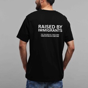 May include: Black t-shirt with the text "RAISED BY IMMIGRANTS" in white block letters. Below the text is the phrase "THE CHILDREN OF THOSE WHO TURNED NOTHING INTO SOMETHING" in smaller white letters.