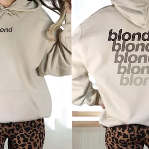 May include: A beige hoodie with the word "blond" printed on the front and back. The back of the hoodie features the word "blond" repeated in a gradient of gray tones. The hoodie has a front pocket and a hood with a drawstring.