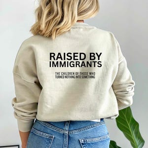 May include: A beige sweatshirt with the text "RAISED BY IMMIGRANTS" in black block letters. Below the text is the phrase "THE CHILDREN OF THOSE WHO TURNED NOTHING INTO SOMETHING". The sweatshirt is worn with blue jeans.