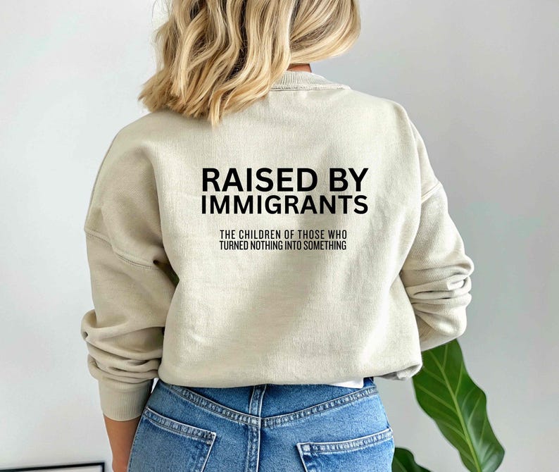 May include: A beige sweatshirt with the text "RAISED BY IMMIGRANTS" in black block letters. Below the text is the phrase "THE CHILDREN OF THOSE WHO TURNED NOTHING INTO SOMETHING". The sweatshirt is worn with blue jeans.