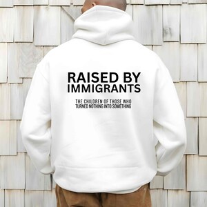 May include: White hoodie with the text "RAISED BY IMMIGRANTS" in bold black letters. Below the text is the phrase "THE CHILDREN OF THOSE WHO TURNED NOTHING INTO SOMETHING" in a smaller font.
