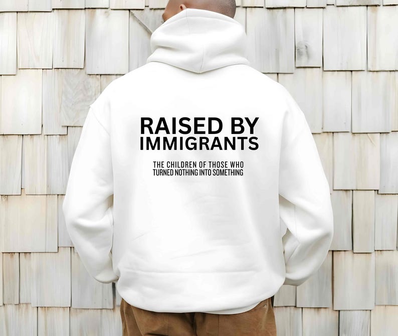 May include: White hoodie with the text "RAISED BY IMMIGRANTS" in bold black letters. Below the text is the phrase "THE CHILDREN OF THOSE WHO TURNED NOTHING INTO SOMETHING" in a smaller font.