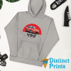 Könnte beinhalten: Ein grauer Hoodie mit einer roten und weißen Illustration eines roten Sportwagens mit dem japanischen Wort "マツダ" unter dem Auto.