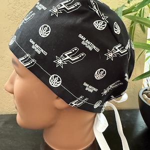 May include: Black surgical scrub cap featuring the San Antonio Spurs logo and text. The cap is patterned with the team's logo and the words "San Antonio Spurs" in white. It has white ties for securing it.