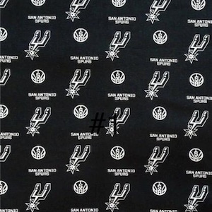 K&ouml;nnte beinhalten: Schwarzer Stoff mit einem sich wiederholenden Muster des Logos und Textes der San Antonio Spurs. Das Design umfasst das Teamlogo, einen stilisierten Sporn mit einem Stern und die Worte "SAN ANTONIO SPURS" in Wei&szlig;.